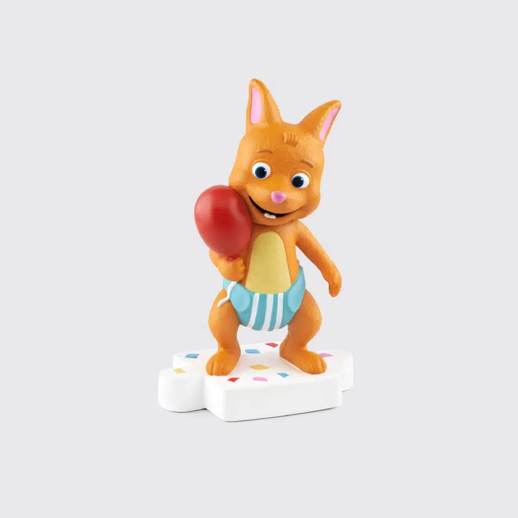 Tonie figure is an orange cat in a blue diaper holding a red balloon and standing on a white puzzle piece platform with sprinkles on it