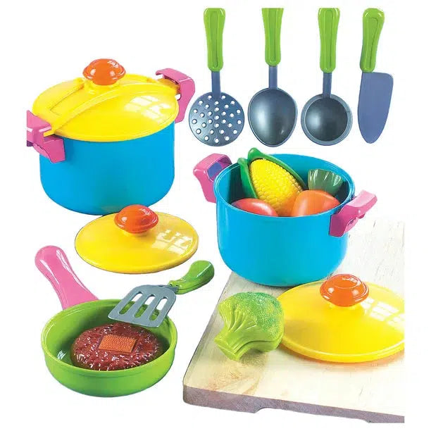 Shows all of the included pieces outside of the packaging. It includes two pots with lids, a frying pan with lid, 5 different cooking utensils, and various food items like corn, tomatoes, carrots, broccoli, and a meat patty.