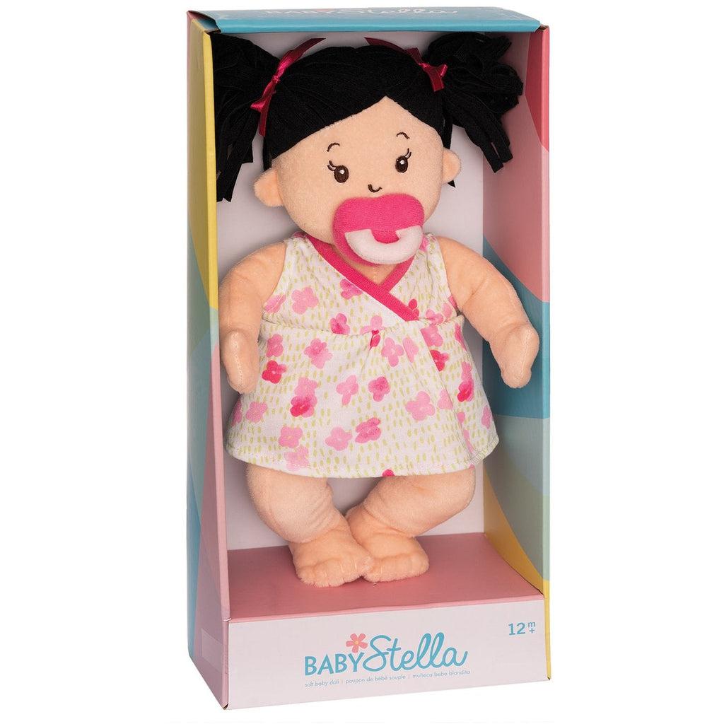 A black haired doll is in a box that reads: "baby stella" at the bottom. The doll has two black pigtails, a pacifier, and a white dress with pink and red flower patter.
