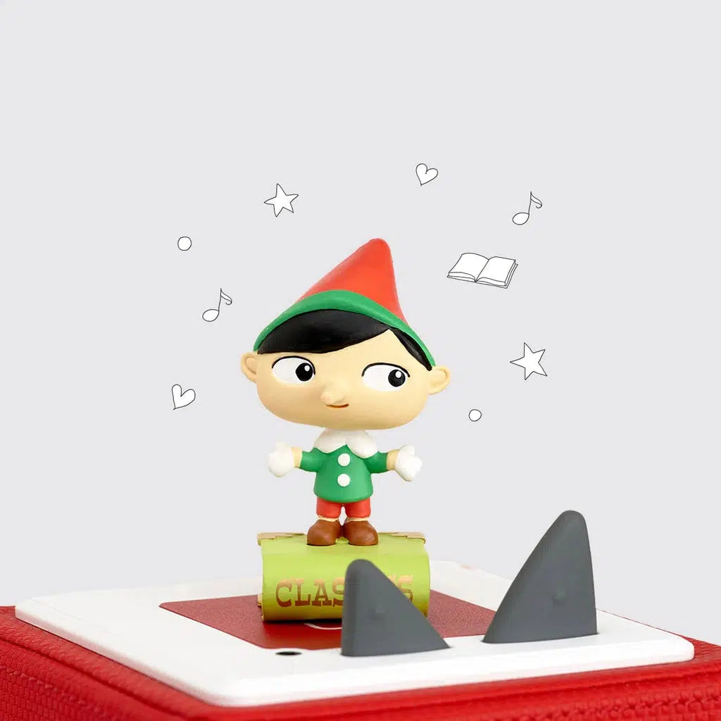 The tonie figure is shown on a toniebox. The figure is a cartoonish pinochio with a wide head. A pointed red hat and green coat standing on a book with "Classics" written on the spine.