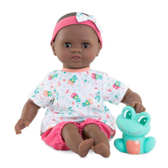 baby doll and frog are shown out of the box. The baby doll is wearing a pink headband with white bow, pink shorts, a white shirt patterned with frogs on lilypads, and is sitting up on it's own