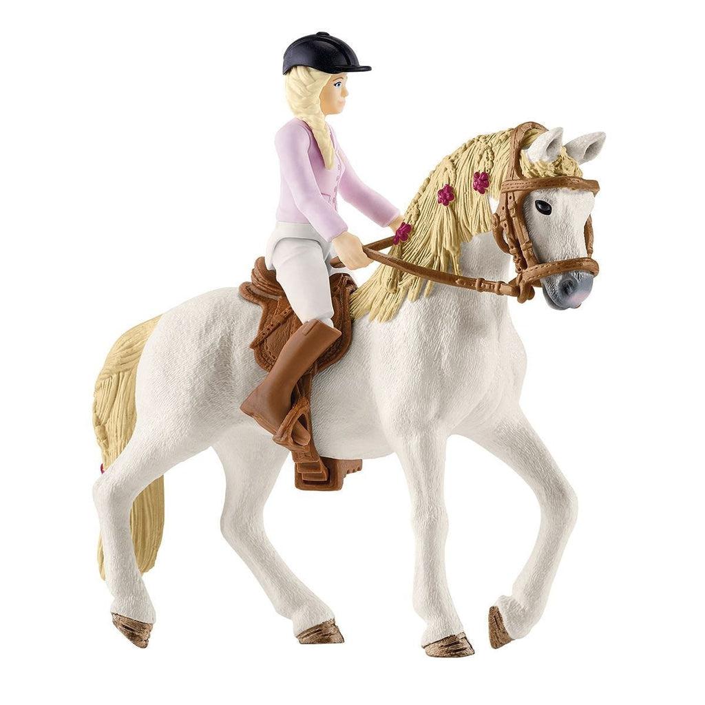Close up of the rider on her horse. The rider is a blonde wearing riding gear and the horse is white with a blonde mane that has flowers woven in. The horse comes with a saddle and bridle.
