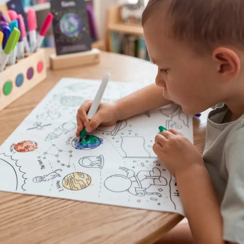 A child colors a space-themed reusable mat at a desk, surrounded by markers and drawing supplies, inspired by creative drawing prompts.