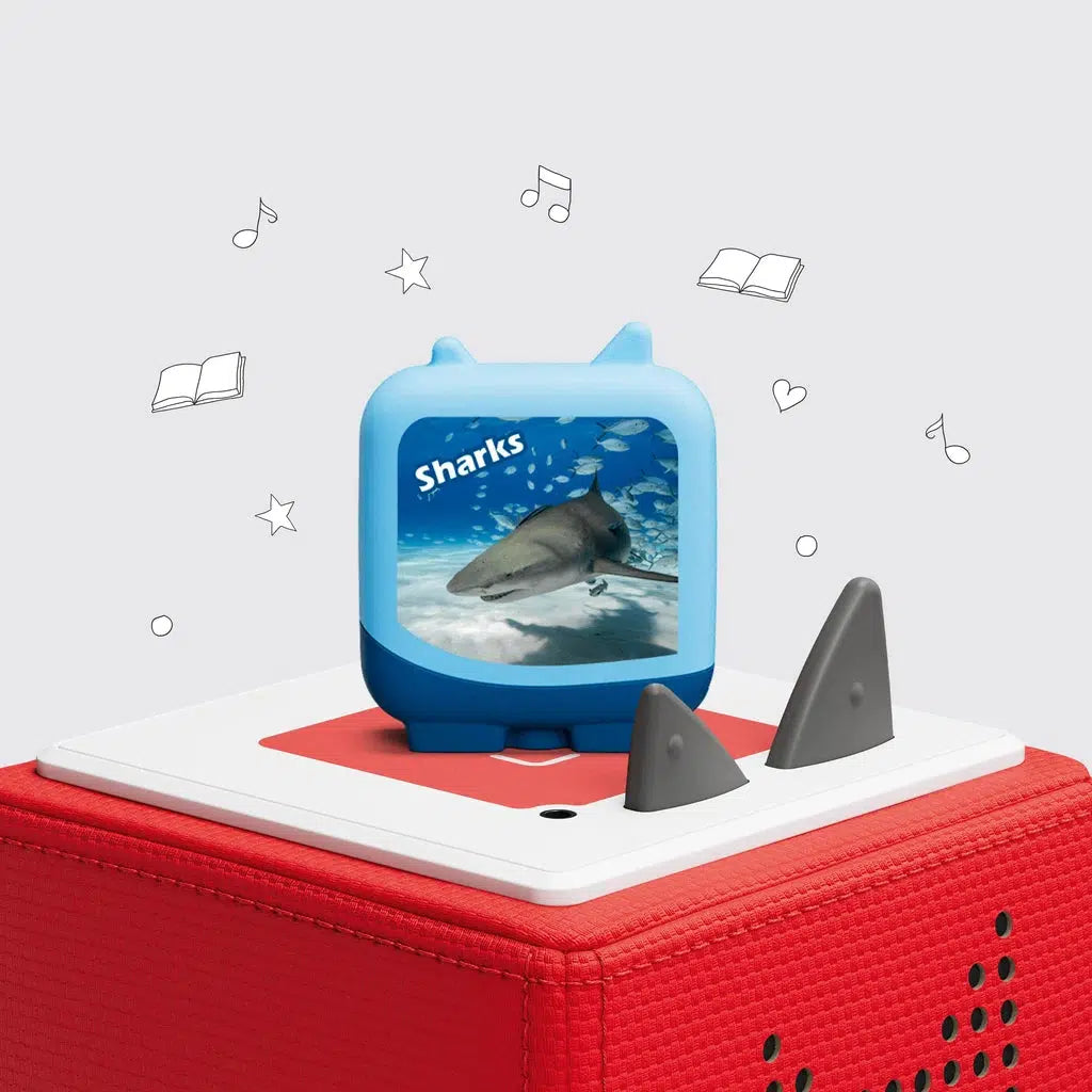 One of the 4 included clever tonies is on a toniebox. This one is a blue square with feet and tonie ears. The front has a picture of a shark with the label "sharks"