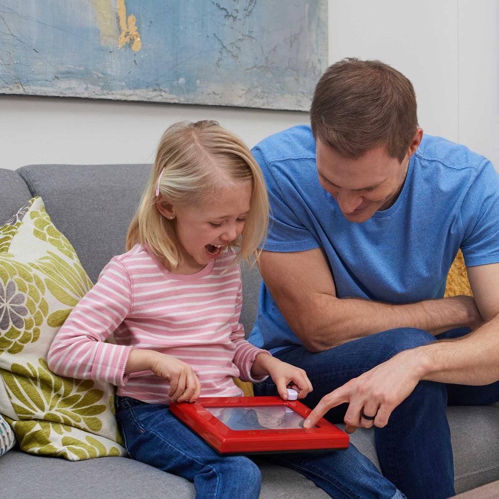 Scene of a father teaching his smiling daughter how to use the Etch A Sketch.
