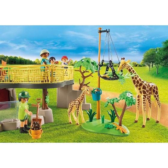 this picture shows the firaffes grazing food on a tray that has been lifted high up. a zookeeper is in the pen making sure the giraffes are healthy and happy. 