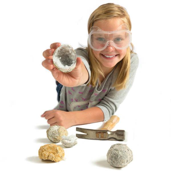 image shows a girl holding up a geode while smiling and wearing safety glasses to not get rock shards in her eye while discovering crystals. 