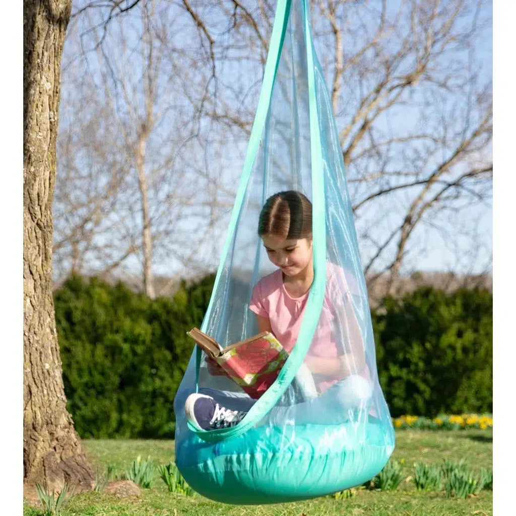 this image shows a girl sitting cross legged in the pod while reading a book. the pod is hanging on a tree branch outside. 