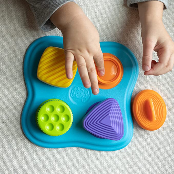 Image of the toy outside of the packaging. It is a blue base with four differently colored shapes. It includes a yellow square, an orange oval, a purple triangle, and a green circle.