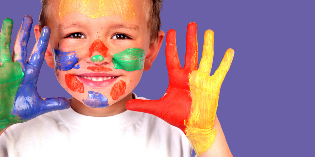 a child is smiling and facing the camera with their hands help up next to their face. Their face and hands are covered with a large amount of paint in various colors.
