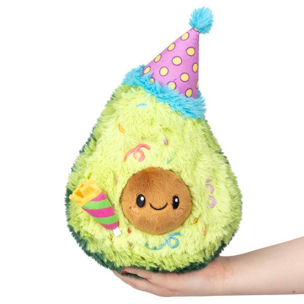 A plush toy of a birthday avocado wearing a party hat, held in a person's hand, with a smiling face and a colorful design.