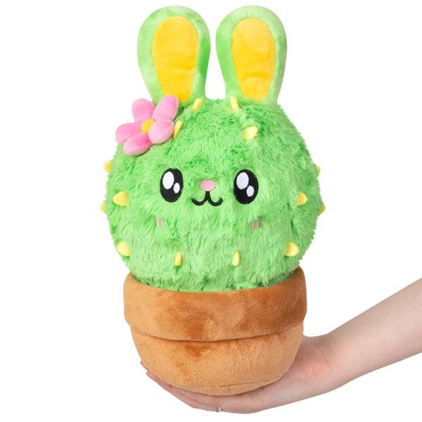 A hand holding a plush toy of a green Bunny Cactus with bunny ears and a flower, in a brown pot.
