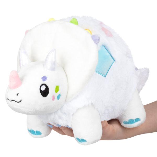 Image of the Mini Opalceratops squishable. It is a completely white dinosaur except for the multicolored pastel embroidered geometric shapes on its face and its sides.
