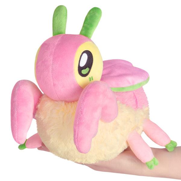 A plush toy  of a cartoon Orchid Mantis, is a whimsical, pink and yellow bug with green antennae and feet, resting on a human hand.