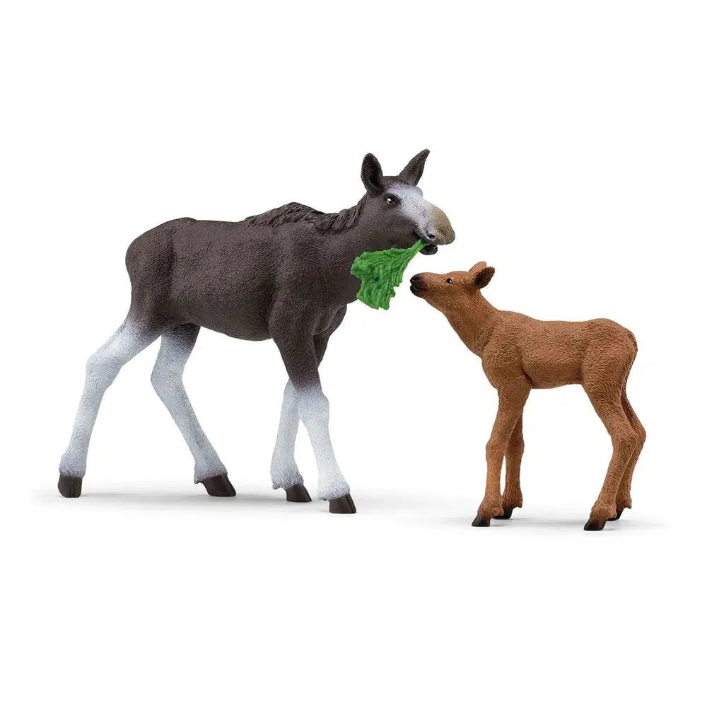 Image of the Moose Family figurine set. It comes with a mother moose and a baby moose. The mom moose is dark grey with white legs and nose and it is holding a leafy branch in its mouth. The baby moose is smaller and is brown. Neither of the animals have antlers.