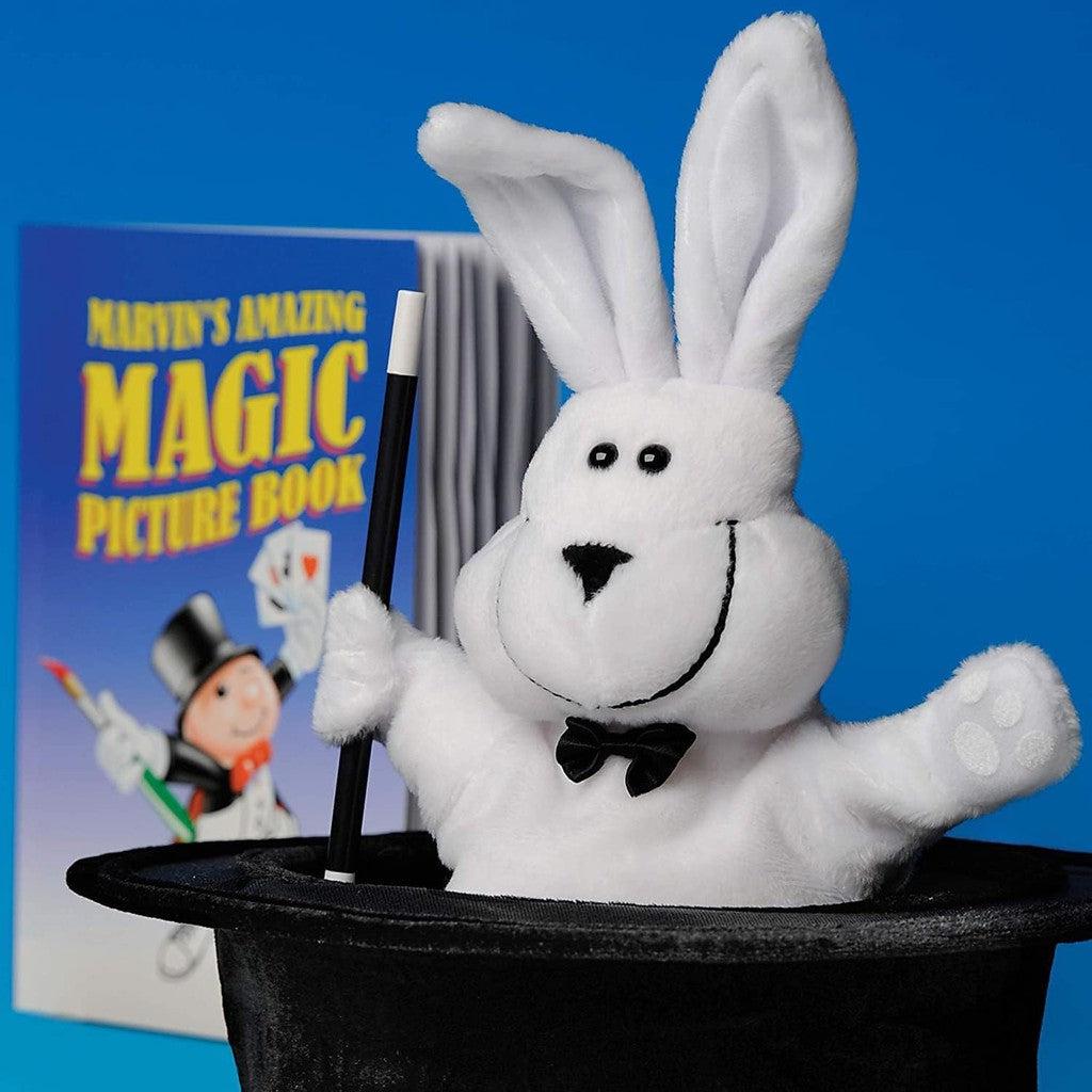 this image shows the stuffed rabbit holding a wand in a hat there is a picture book behind the rabbit