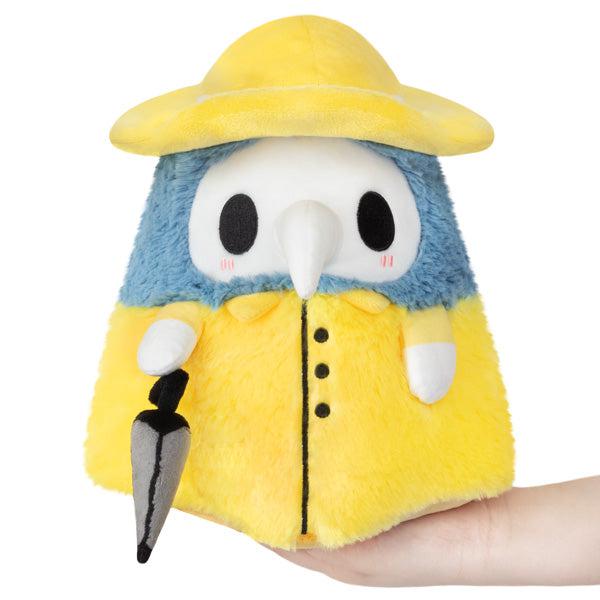 A Mini Squishable Rainy Plague Doctor plush toy resembling a bird dressed in a yellow raincoat and hat, holding a black umbrella, cradled in a human hand against a white background