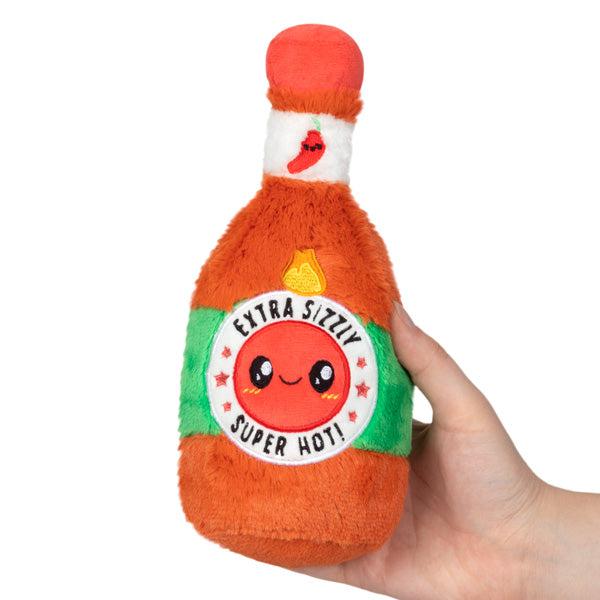 A plush toy resembling a Hot Sauce bottle held in a hand, featuring a smiling face and labeled "extra sizzly super hot!" with red and green colors.