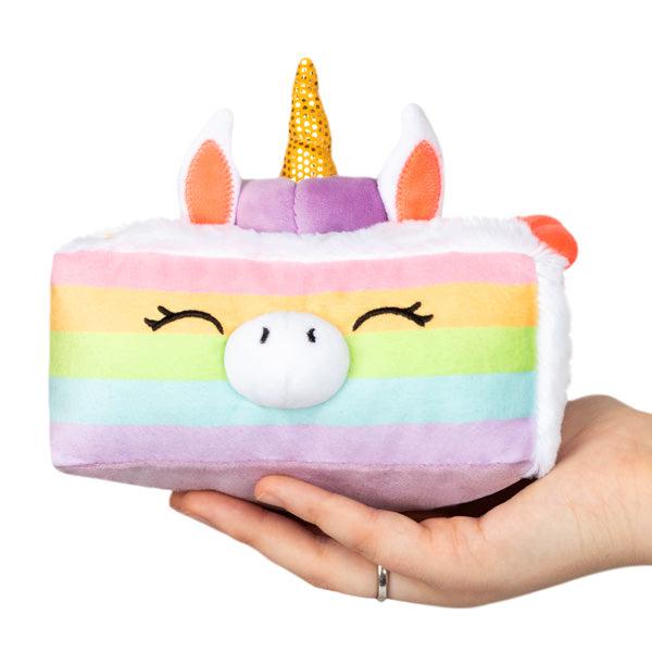 Hands holding a small squishable plush unicorn with a rainbow design, closed eyes, and a gold horn.