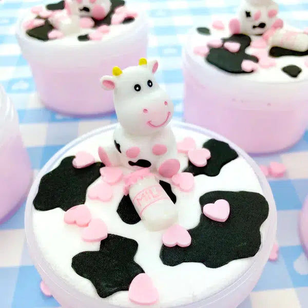 Image of the opened slime. It is a dual colored slime with a pink bottom and a white top. On the top is a black and white cow print design. The slime comes with light pink heart charms and a clay cow topper.