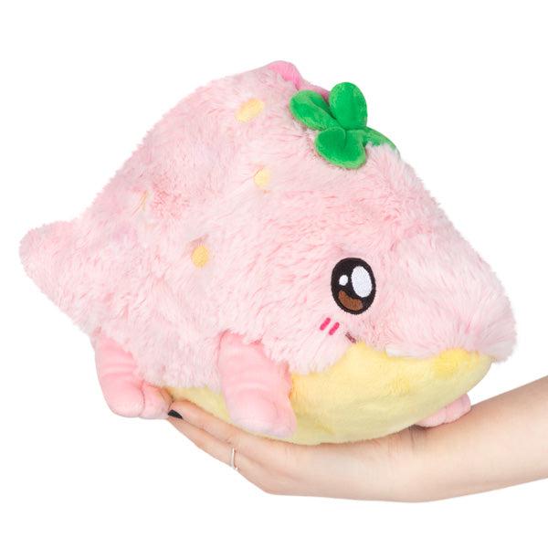 A hand holding a soft, pink Mini Squishable Strawberry Crocodile plush toy shaped like a stylized animal with large eyes and a green detail on top.