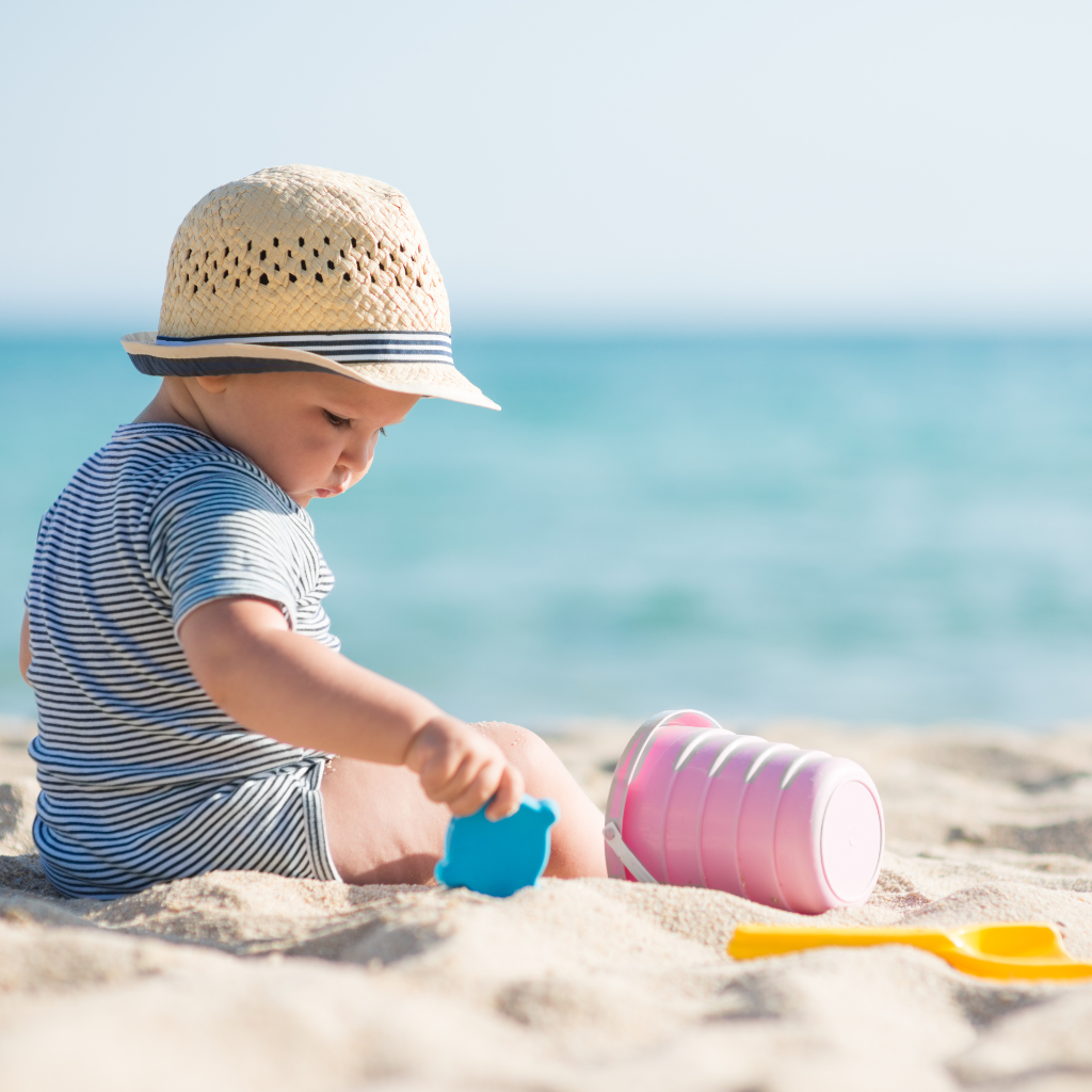 A baby is on a beach wearing a sun hat. The baby is holding a plastic sand scoop and there's a small plastic shovel and bucket next to it