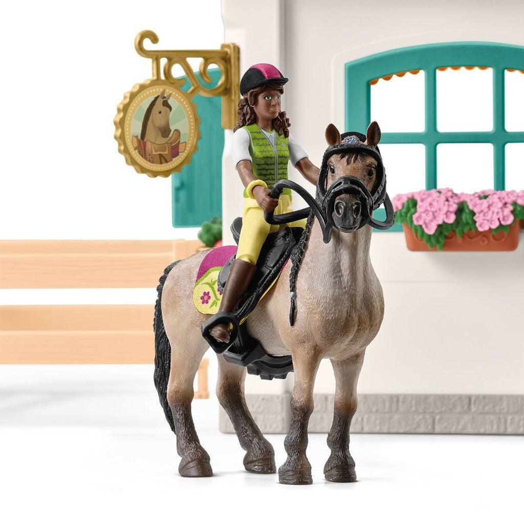 The girl figurine is sitting on the back of the horse and holding the reigns. The corner of the building behind them has a sign depicting a horse behind a fence with a saddle draped over the fence