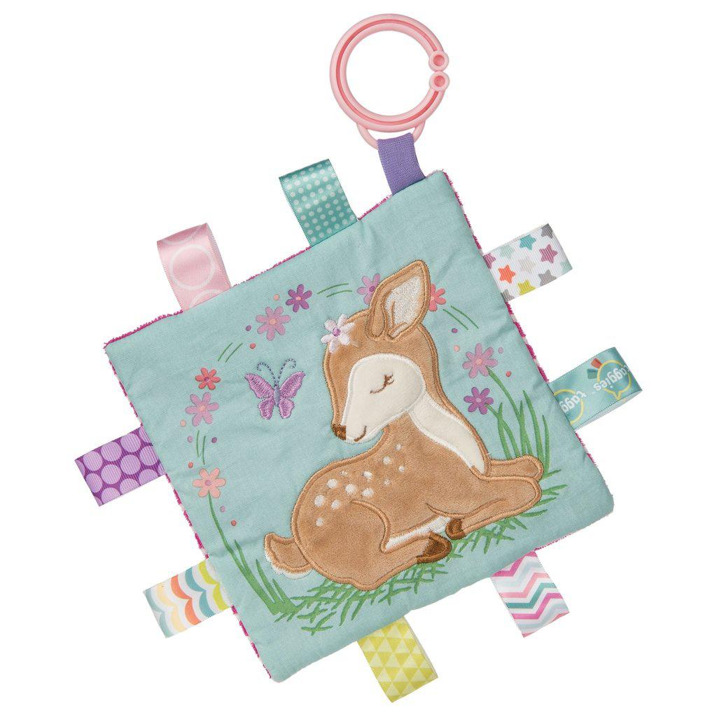Image of the Taggies Crinkle Me Flora Fawn. On the front is an embroidered fawn resting on her knees surrounded by flowers and a butterfly. On the edges are ribbon tags made from fabrics that fit the color theme.