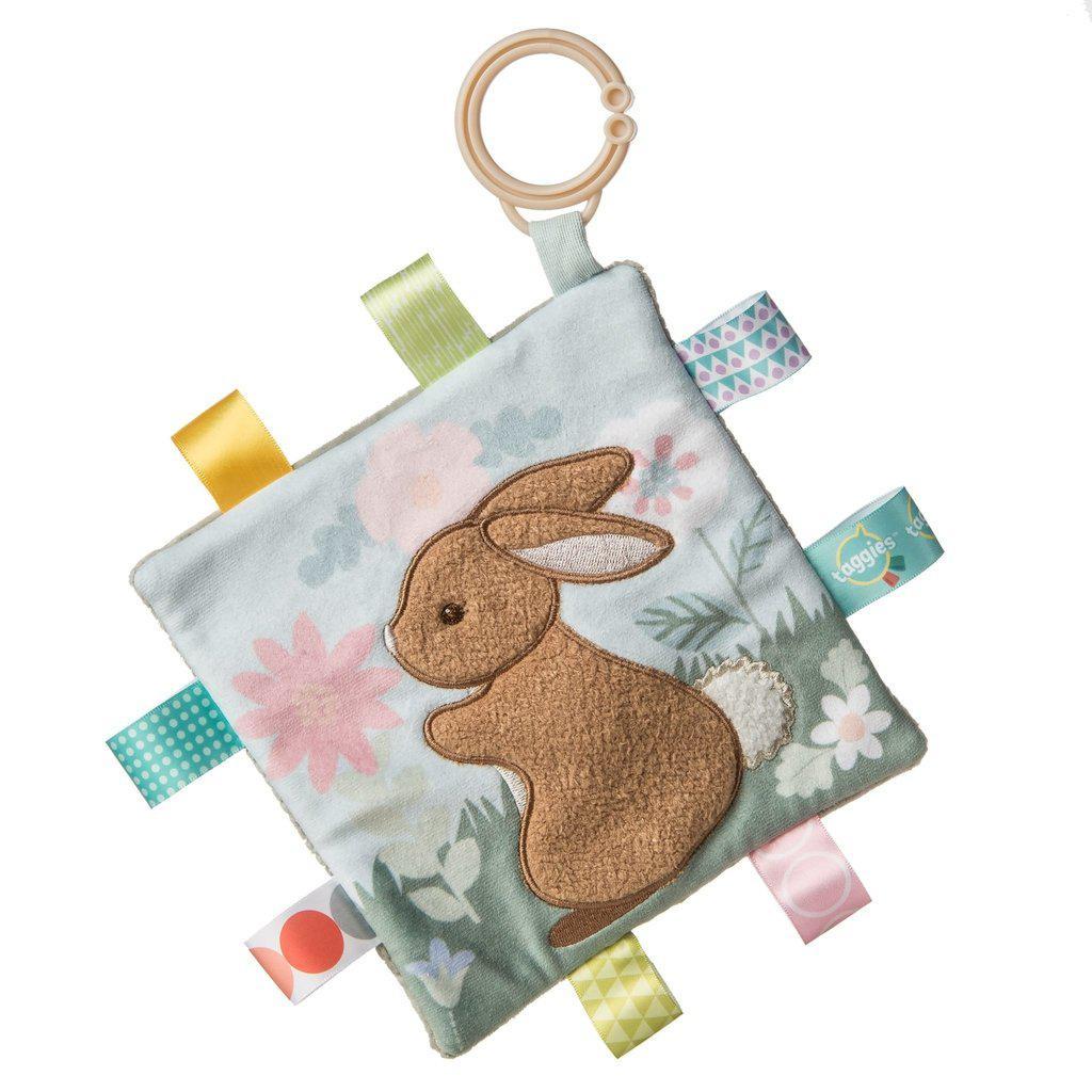 Image of the Taggies Crinkle Me Harmony Bunny. On the front is an embroidered bunny with soft fur in a field of flowers. On the edges are ribbon tags made from fabrics that fit the color theme.