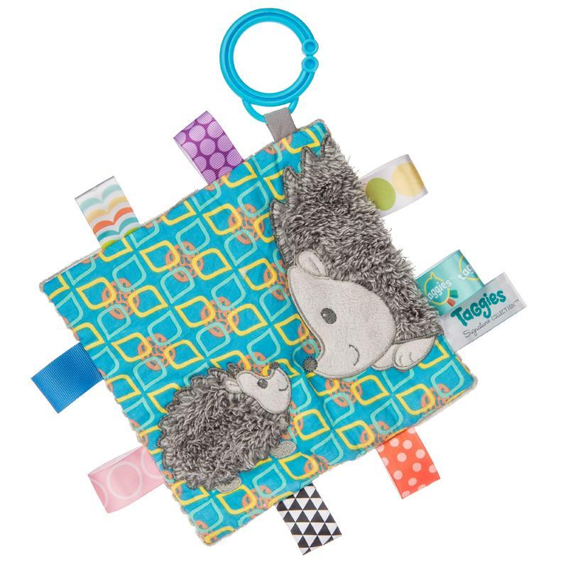 Image of the Taggies Crinkle Me Heather Hedgehog. On the front is are two embroidered hedgehogs (one mom and one baby) touching noses with fluffy spines. On the edges are ribbon tags made from fabrics that fit the color theme.