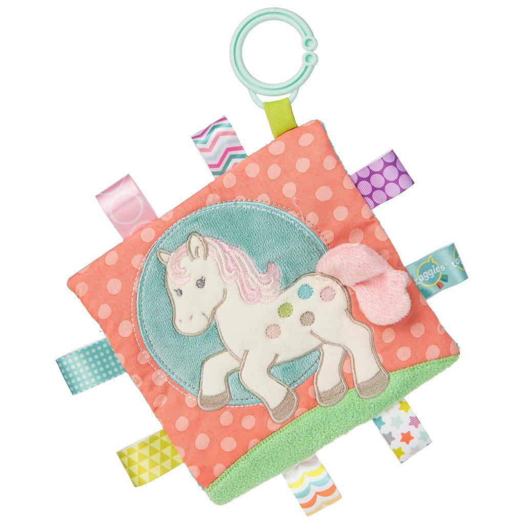 Image of the Taggies Crinkle Me Painted Pony. On the front is an embroidered pink and cream pony with different colored spots on her flank. Her tail is soft and comes off the picture. On the edges are ribbon tags made from fabrics that fit the color theme.