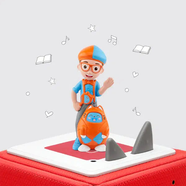 The blippi tonie is a boy with suspenders and an orange and blue newsboy cap with a robot cat by his feet.