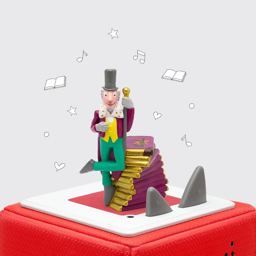 Tonie figure is on top of a tonie box. The figure is of willie wonka with grey sideburns and a tall grey tophat, a maroon coat, yellow vest, and green pants next to a pile of giant chocolate bars
