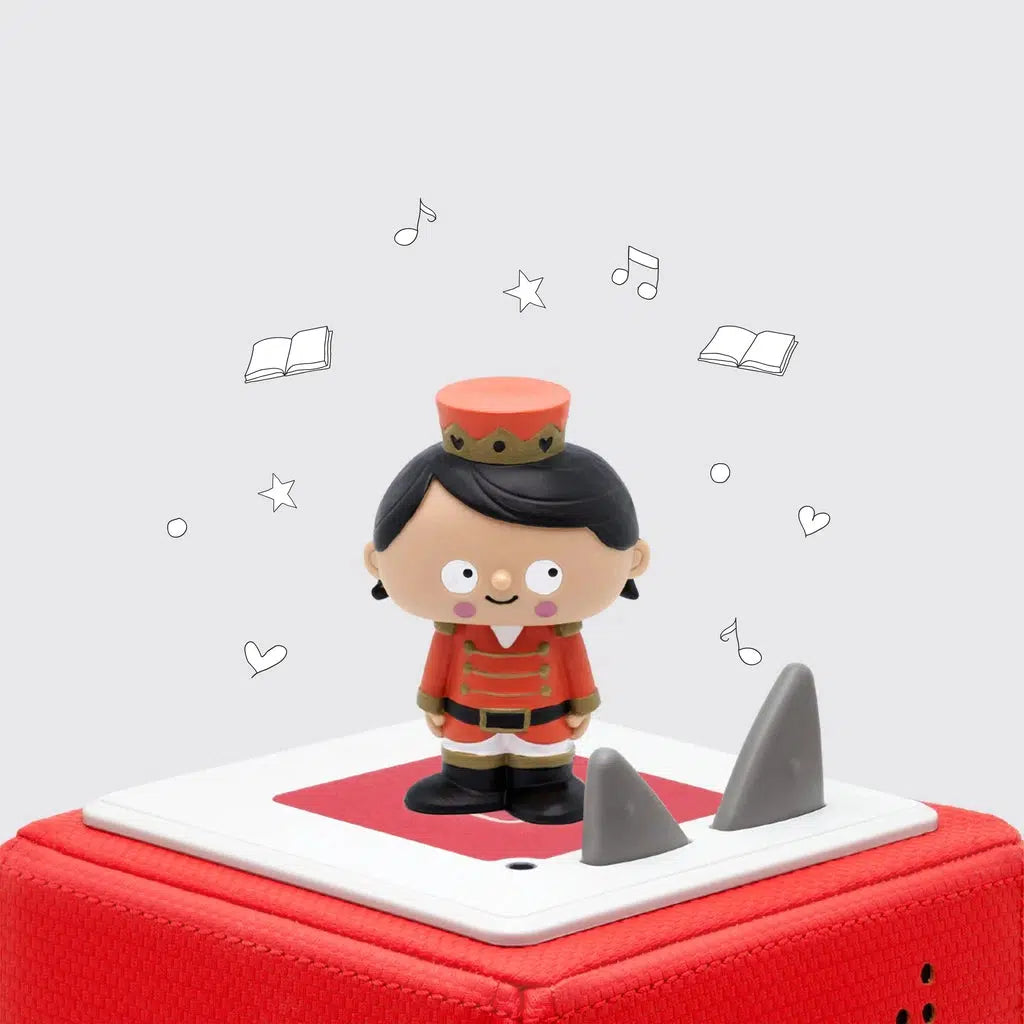 The tonie figure is shown on a toniebox. The figure is a nutcracker boy, wearing a red coat with gold trim, black boots and belt, and a round red hat with gold trim at the base.