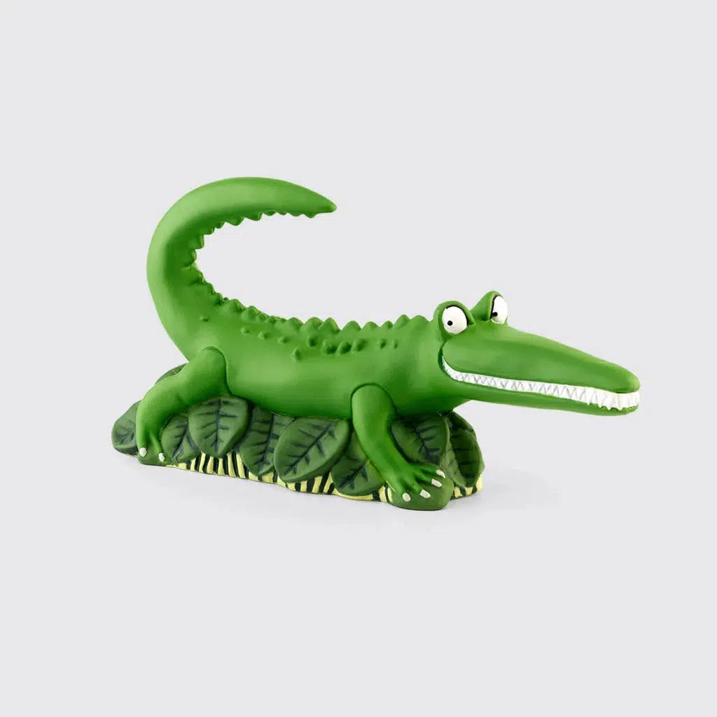 The tonie figure is a green crocodile with a toothy suspicious grin sitting on a pile of large leaves