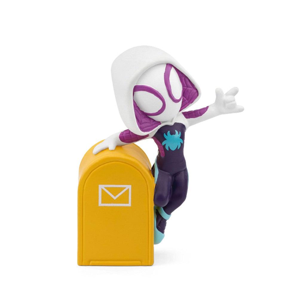 Tonie figure is gwen stacey's spiderwoman, a white costume with purple lining in the hood and around the eyes with a black and blue jumpsuit over the rest of the body. She's standing on the side of a yellow mail deposit box