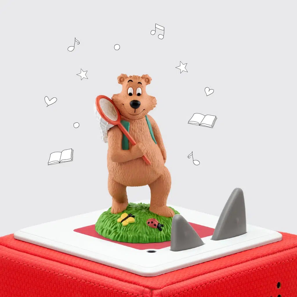 The tonie figure is shown on a toniebox. The figure is a cartoonish bear on two legs holding a red bug net and wearing a green vest standing a a patch of grass with a butterfly and a ladybug.