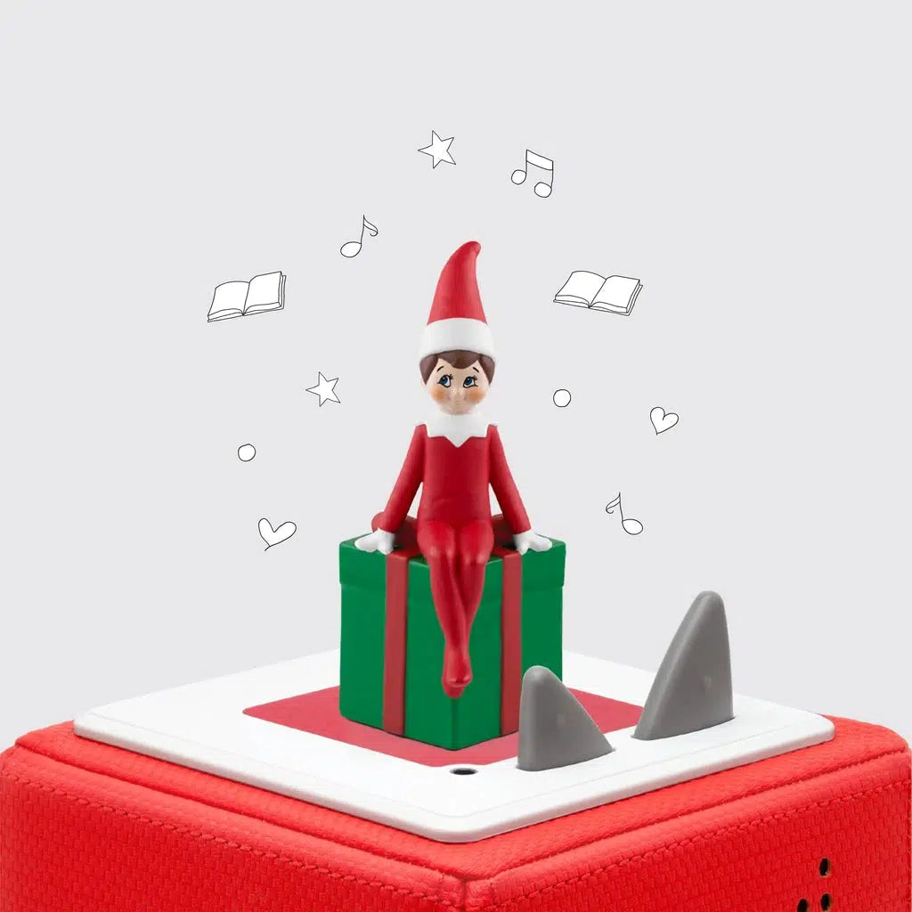 The tonie figure is shown on top of a toniebox. The figure is an "elf on the shelf" sitting on the edge of a green gift box with a red bow. 