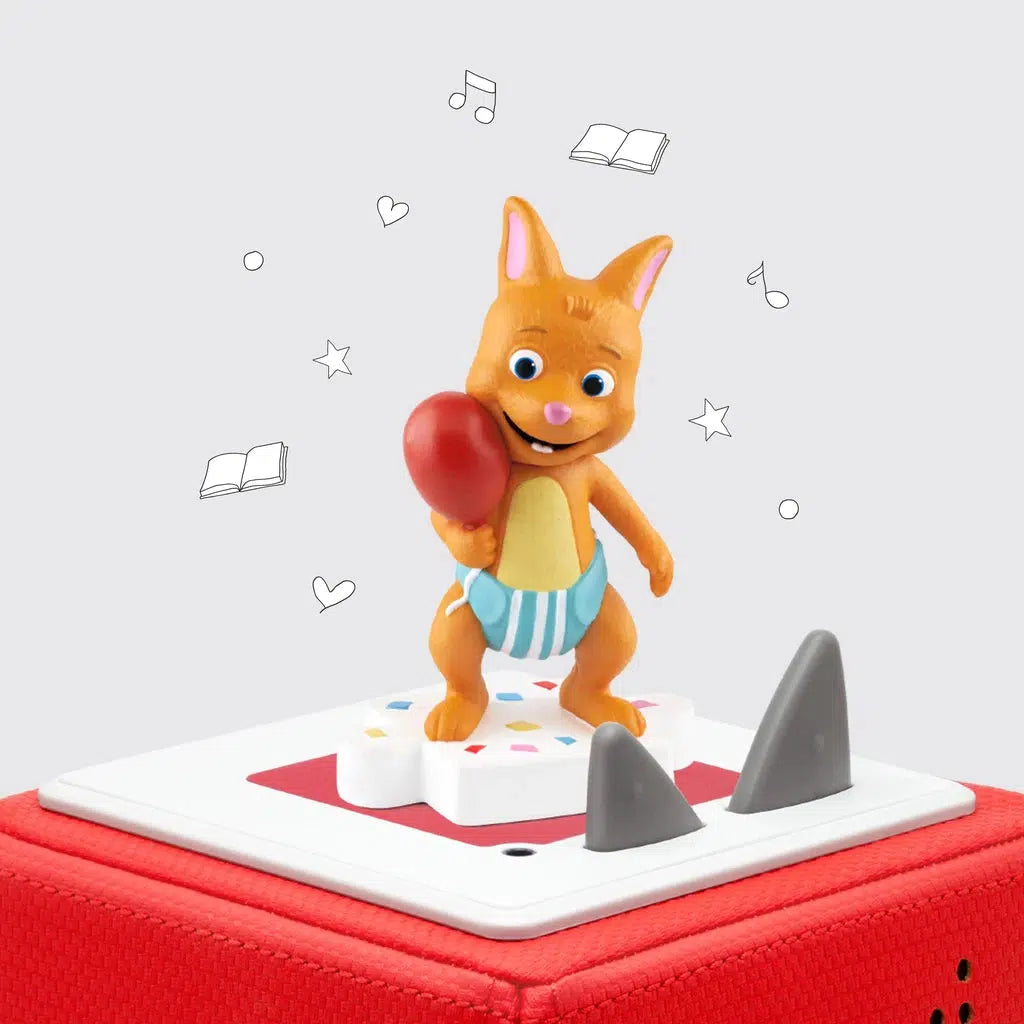 The tonie figure is shown on a toniebox. The figure is an orange rabbit looking cartoon character holding a balloon and wearing a diaper standing atop a base colored like white frosting with sprinkles in it.