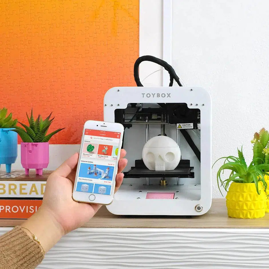 A white 3D printer labeled "TOYBOX" sits on a shelf, printing a white skull-shaped bject. Nearby, a hand holds a smartphone displaying apps and 3D models for printing