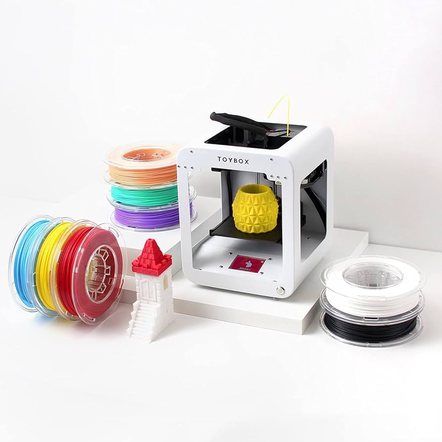 A white 3D printer for kids with the name "Toybox" on it is surrounded by spools of filament in various colors. An orange object is being printed inside the printer