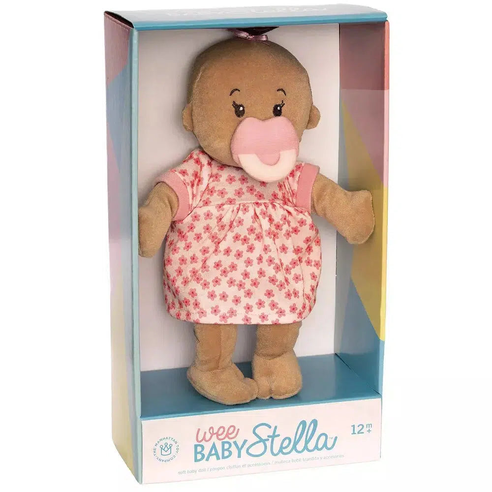 wee baby stella is a soft doll for babies. this image shows wee stella in a doll box with a pink skirt, and pink pacifier. baby stella has brown skin