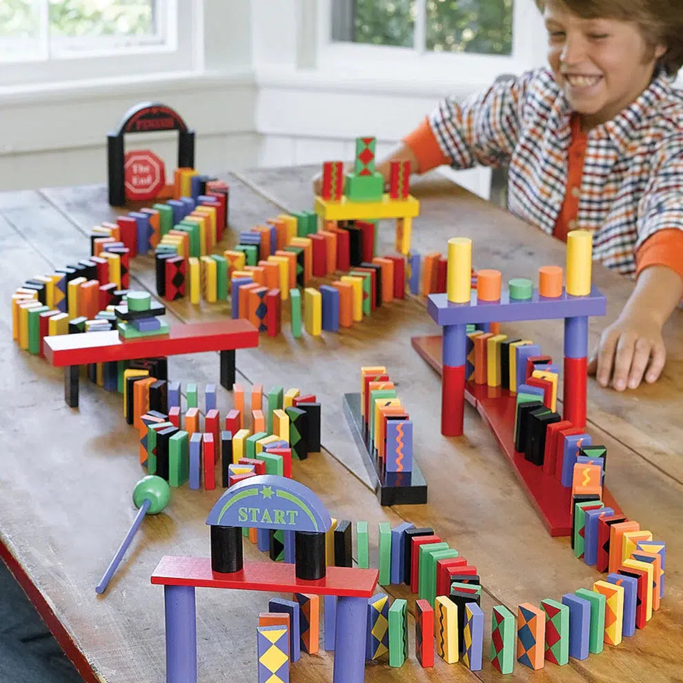 A boy sits at a wooden table smiling at the winding domino track he has set up. The dominoes are varying colors and wind in a serpentine fashion as it travels up and down ramps and around poles and under crossing beams.