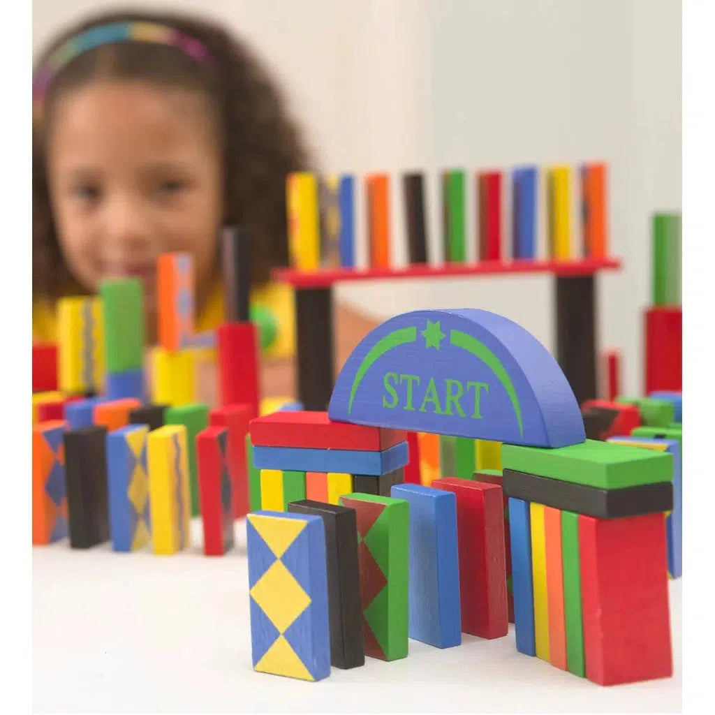 A close up of the start gate of a domino track with an out of focus girl in the background. The start gate is a half circle that spans a gap between two pillars of 4 dominoes vertically lined up and 2 horizontally on top of them.