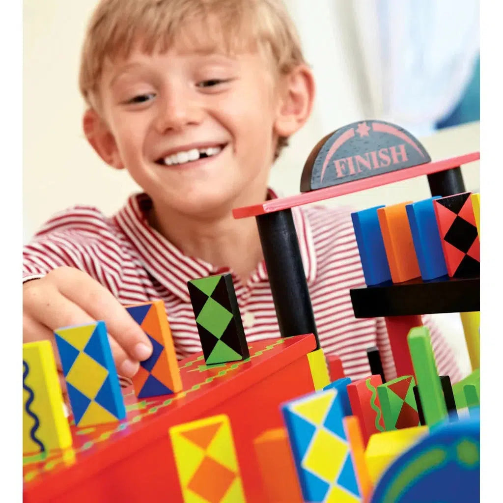 A boy smiles as he places a domino into the middle of a domino track. He is placing it on a raised area and the track can be seen coming in and out of view in the bottom right and the finish gate is in view at the back right in front of the boy.