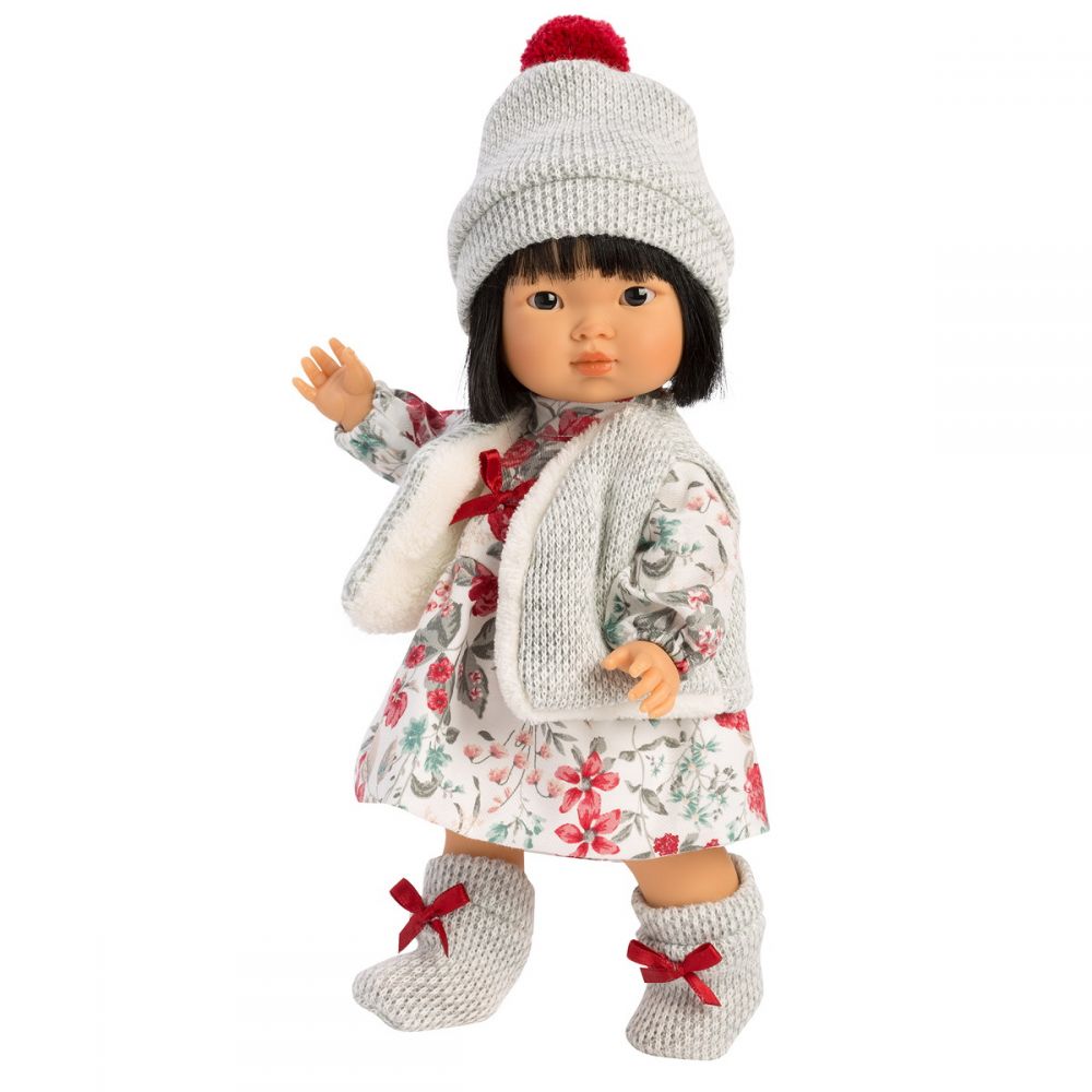 Llorens Aja Doll | This doll has brown eyes and black hair styled into a short bob with straight across bangs. She is wearing a white dress with red and gray floral print. She has on a gray vest with white fur lining secured by a red ribbon. Her hat and socks are made of the same gray material as the vest. The socks have red ribbons details, and the hat has a red pom-pom at the top. 