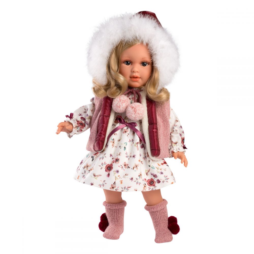 Doll out of package | Doll has long, blonde, curled, hair and blue eyes. She is wearing a white dress with pink floral prints. She has a pink fur lined vest with a hood and red metallic details. She has on pink socks with red pom-poms.