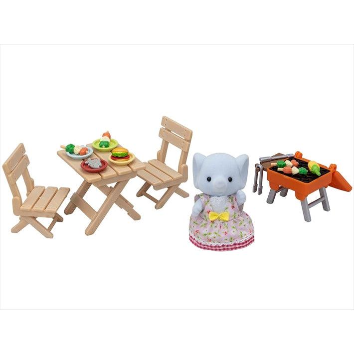 Shows the elephant girl standing in front of the picnic table with food on the plates and the grill with food grilling.