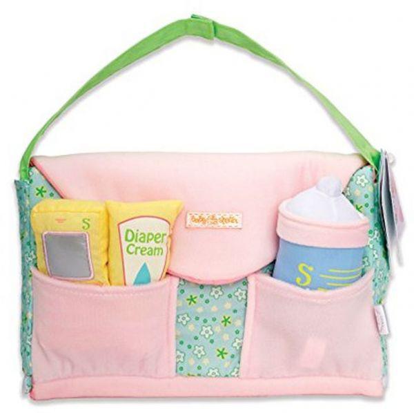 A toy diaper bag is shown. The base is a green pattern with white flowers and dots spread around it. It has two pockets on the front and a flap opening for the inside of the bag on top. There is a toy bottle and fake plush diaper cream in the pockets on the front.