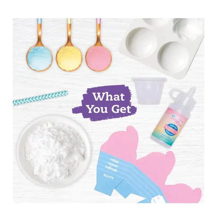 Text in the center reads: What you get. Image shows 3 spoons filled with blue, yellow, and red baking soda, a tray with the various food shapes for molding the bath bombs, a bowl of white citric acid. A mini milkshake cup for shaping, scented glycerin, and an unfolded gift box.